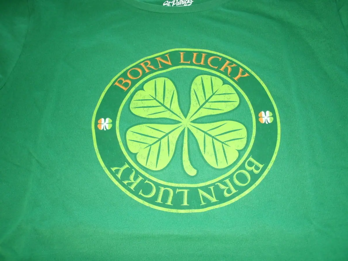 Close-up of a green T-shirt with a circular shamrock graphic and repeated “BORN LUCKY” lettering.