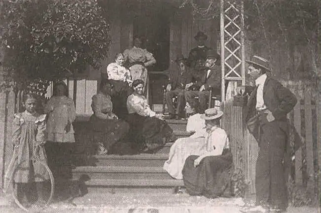A group of adults and children pose on the steps of a wooden porch beside a man in a hat.