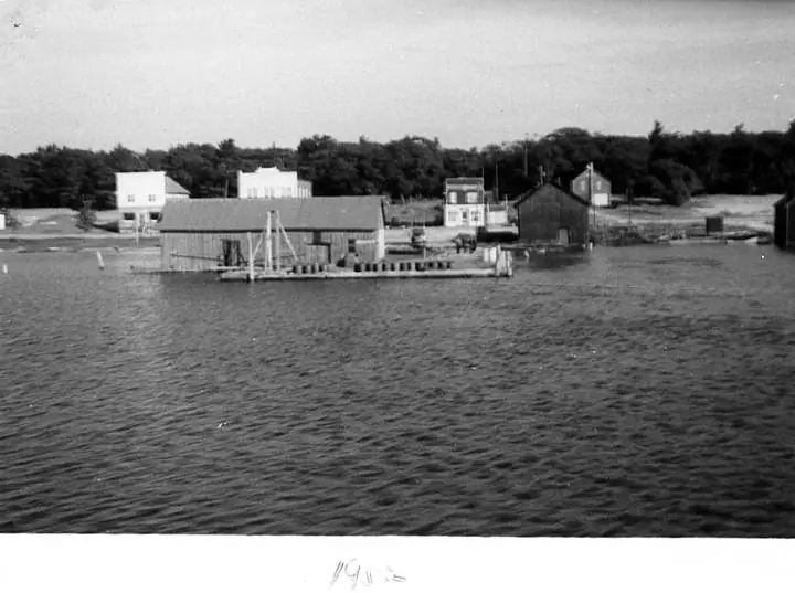 Black-and-white waterfront photograph showing a dock, waterside buildings, and a wooded shoreline.