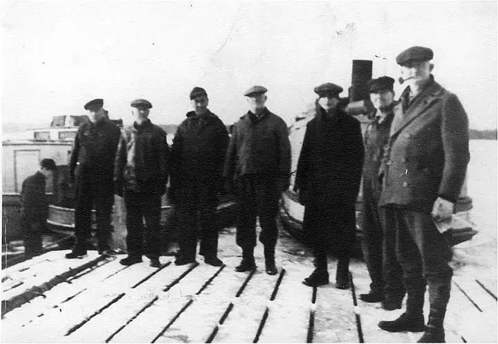 Seven men in heavy clothing stand on a snow-dusted wooden deck beside vessel equipment.