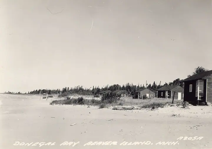 A sandy shoreline with small wooden cabins, dunes, and trees, viewed in a monochrome photograph.