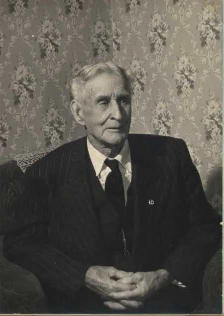 Seated older man in a dark pinstriped suit and tie, posed before floral wallpaper.