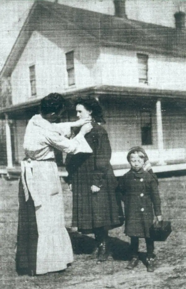 Three females, including a child, stand before a large two-story house as one adjusts another’s coat collar.