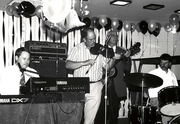 Four musicians perform with keyboards, violin, guitar, and drums beneath balloons and streamers.