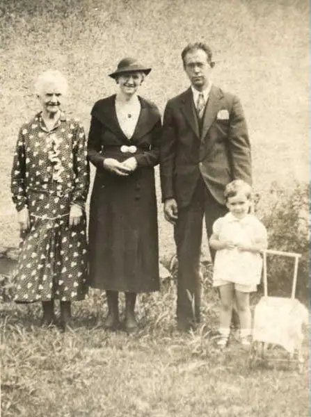 Four people pose outdoors in formal clothing, with a light-colored chair partly visible behind a child.