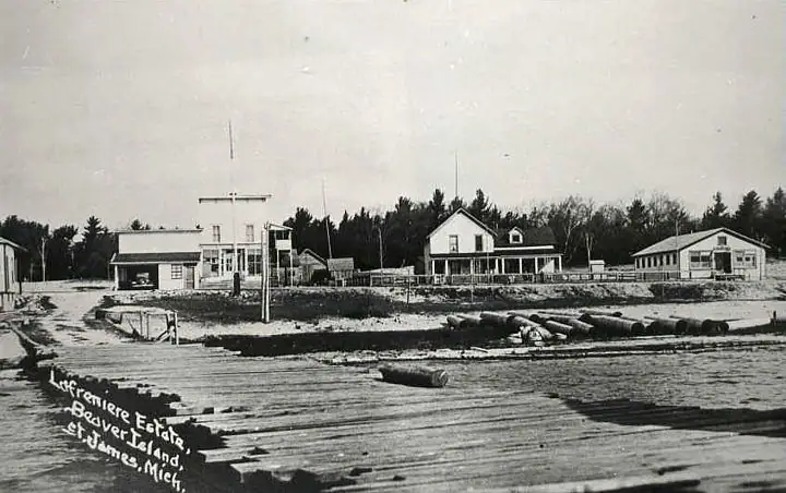 Black-and-white waterfront photograph showing a wooden dock, floating timbers, and several shore-side buildings.