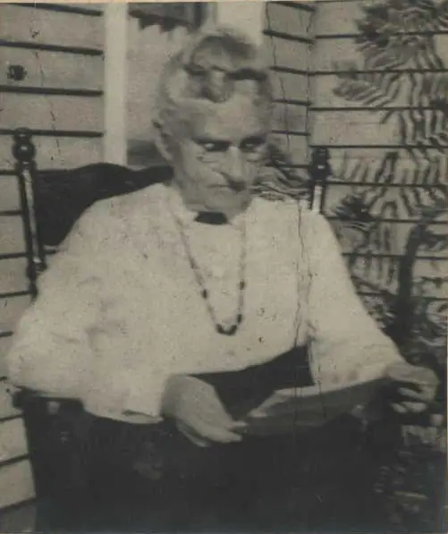 Black-and-white portrait of an older woman seated in a chair, wearing a light blouse and long necklace.
