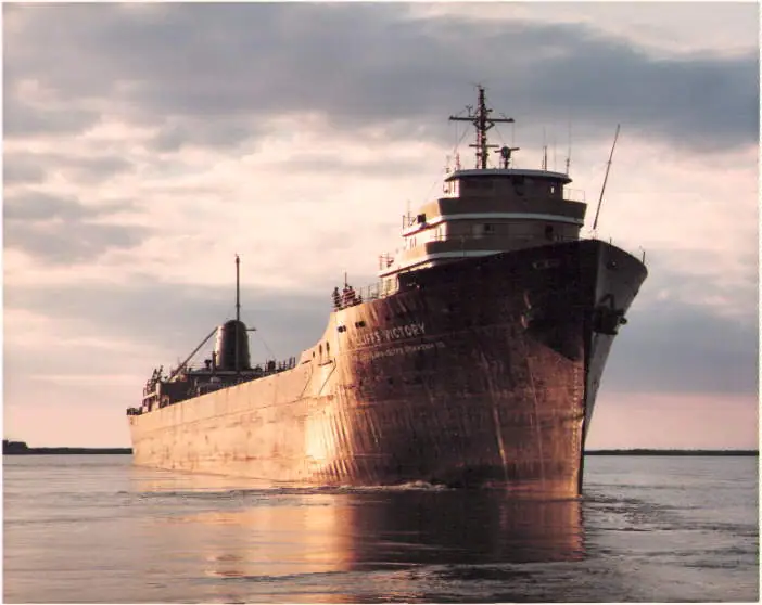 A large dark-hulled ship approaches across calm water beneath dramatic clouds.