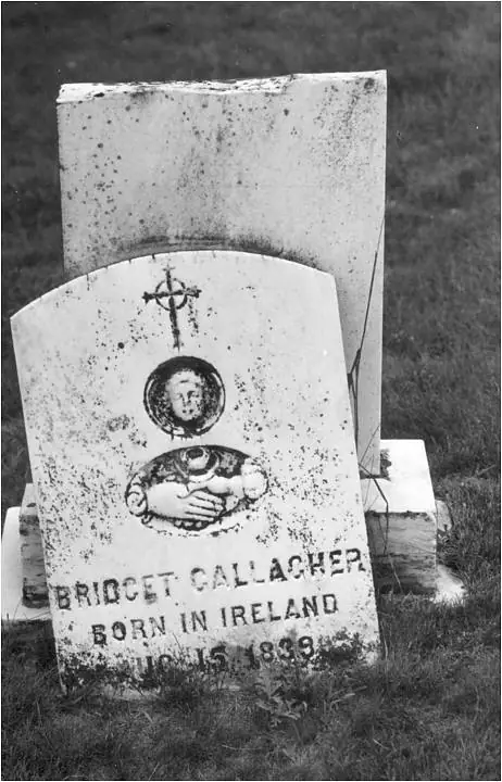 A weathered gravestone with a carved cross, face, and relief of hands standing in grass.