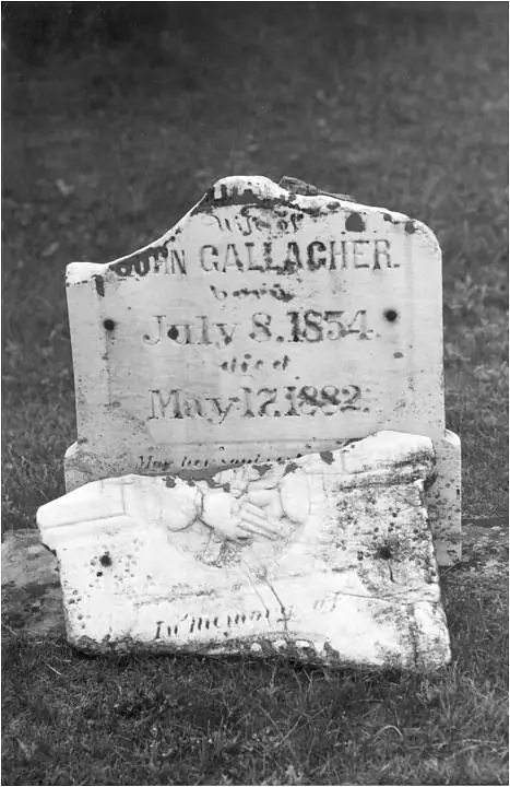 A weathered, broken white grave marker stands in grass, with an inscription and two dates visible.