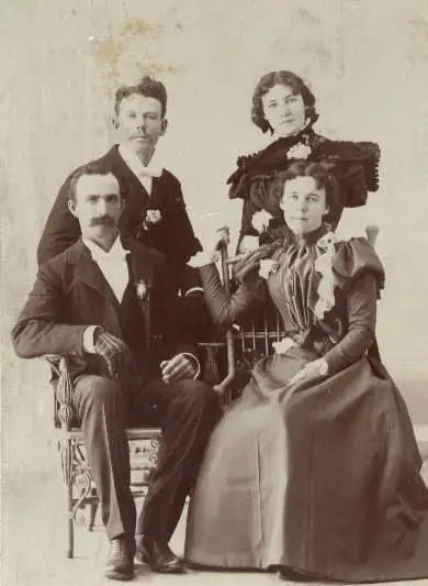 Studio portrait of two seated men and two standing women in formal clothing.