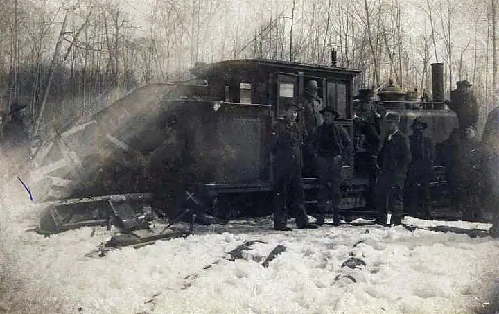 A small steam locomotive and several men stand beside snow-covered railroad tracks in a leafless forest.