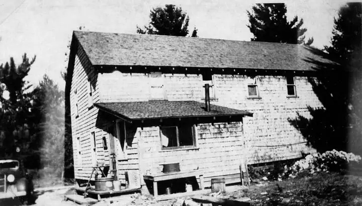 Black-and-white photograph of a weathered wooden building with a broad shingled roof, trees, and a partial vehicle at left.