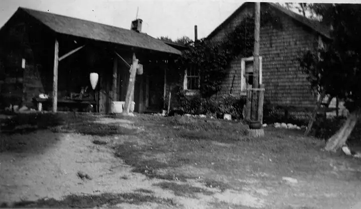 Black-and-white photograph of a shingled wooden house beside an open-sided woodshed and yard.