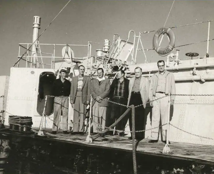 Six men pose on a ship's deck beside railings, pipes, and a lifebuoy marked DETROIT.