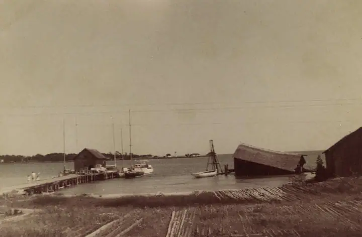 Wooden pier and sailboats beside waterfront sheds, with timber stacked along the foreground.