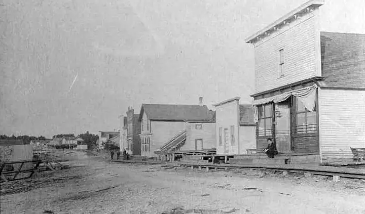 Black-and-white street scene with wooden buildings, an unpaved road, tracks, and several distant people.