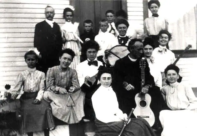 Black-and-white group portrait of adults and children with string instruments outside a wood-sided building.