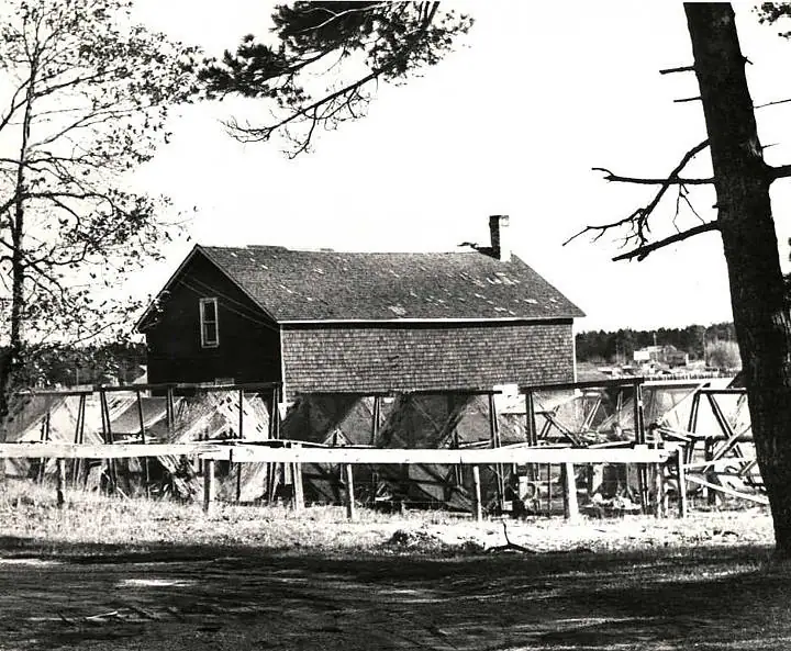 Black-and-white photograph of a shingled wooden fishing shed raised above a timber framework beside the waterfront.