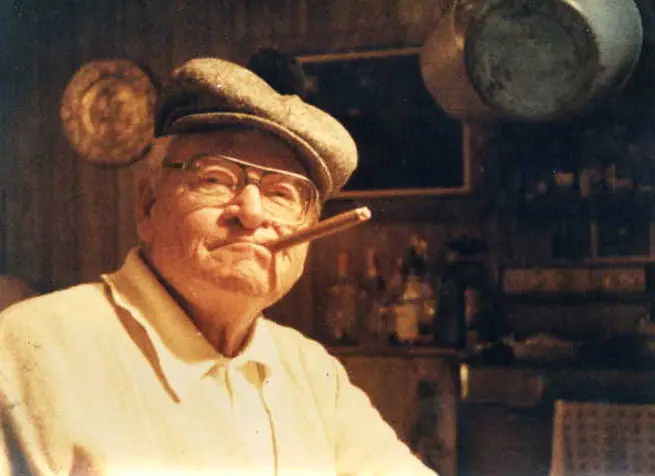 Portrait of an older man in a flat cap and glasses, holding a pipe in his mouth inside a dim room.
