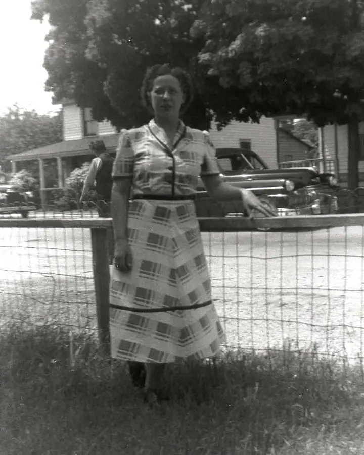 A woman in a patterned dress stands beside a wire fence in front of a house and parked automobile.