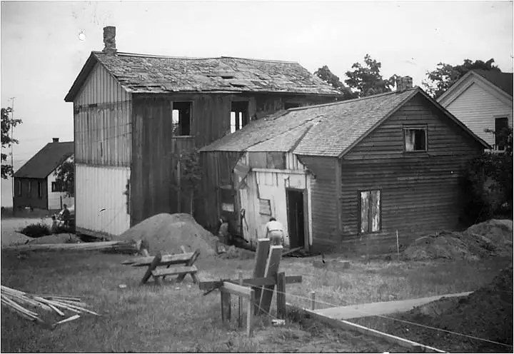 Black-and-white photograph of wooden buildings undergoing construction or restoration, with materials and two people in the foreground.