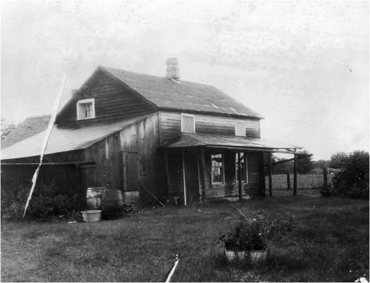 Black-and-white photograph of a weathered wooden farm building with a porch, chimney, barrels, and a potted plant.