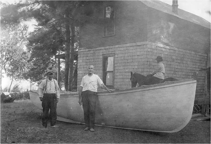 Three men pose beside a large wooden boat in front of a shingled house.
