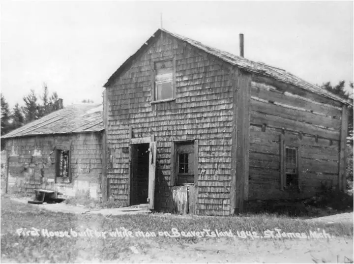 Black-and-white photograph of a weathered wooden house and adjoining outbuilding with shingle siding.