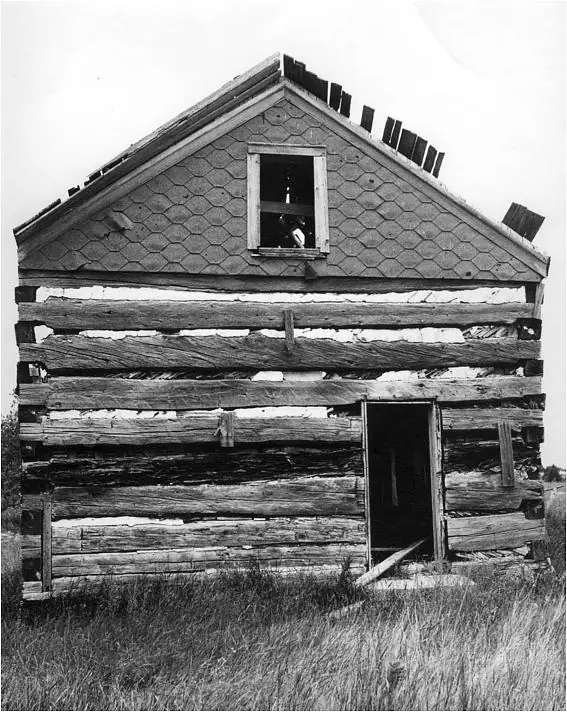 Black-and-white photograph of a weathered log building with a gabled, shingled upper facade and open doorway.