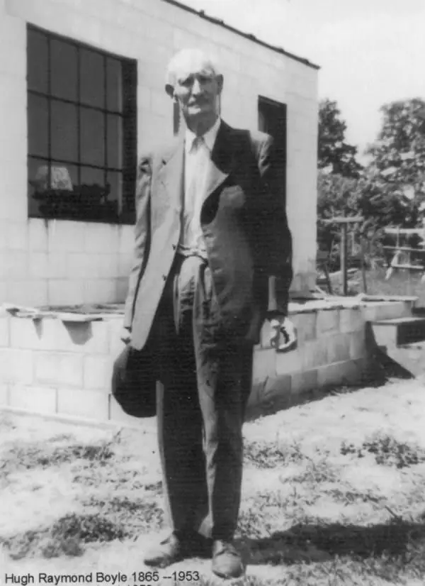 Black-and-white portrait of an older man in a suit standing beside a block building.