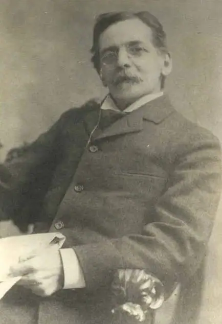 Black-and-white portrait of a mustached man in a suit holding a paper beside a carved chair.