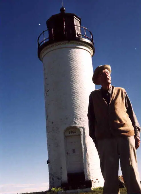 An older man stands beside a tall white cylindrical lighthouse beneath a clear blue sky.