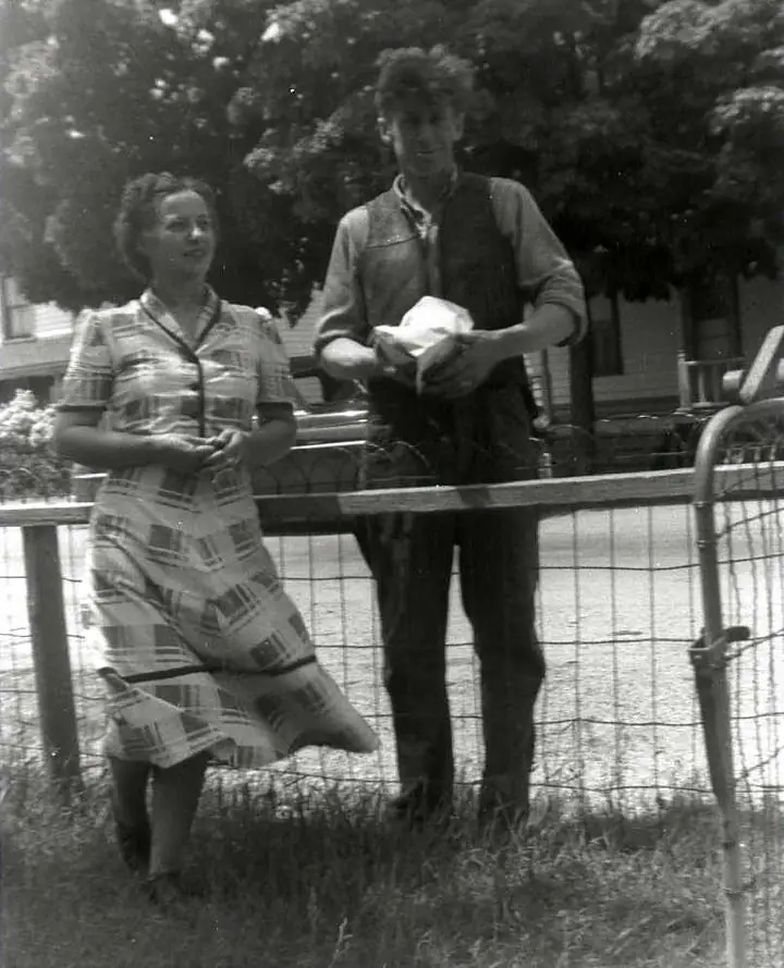 Two adults stand behind a wire fence outdoors, with trees and a house behind them.
