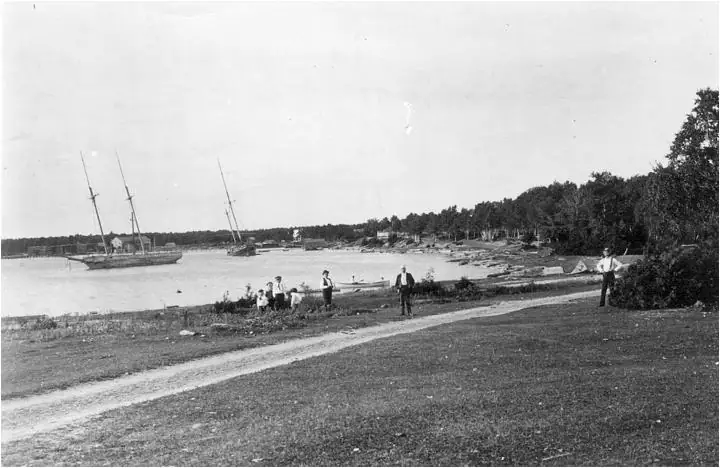 Black-and-white photograph of two schooners near a wooded shoreline with people walking along a path.