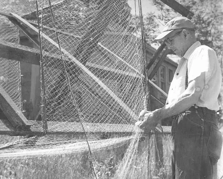 A man repairs a large fishing net stretched across a wooden frame.