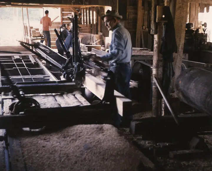 Workers operate large machinery inside a wooden workshop filled with lumber and sawdust.
