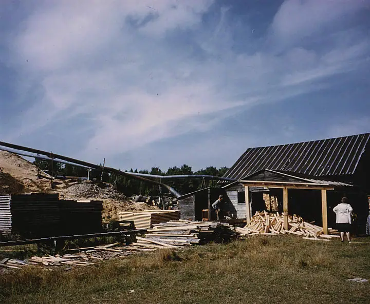 A lumber-processing yard with a conveyor, stacks of boards, a wooden shed, and two people under a cloudy sky.