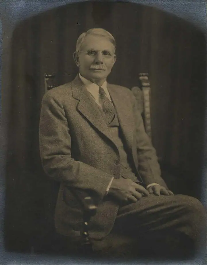 Sepia portrait of a mustached man wearing glasses and a suit, seated in a wooden chair.