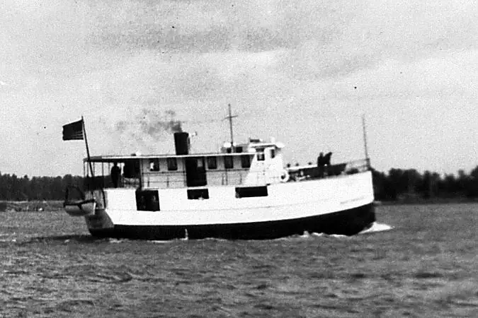 Black-and-white photograph of a small ferry traveling across choppy water near a wooded shoreline.