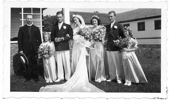 A wedding party of seven people stands on grass outside wood-sided buildings, with bouquets and a bridal gown train.