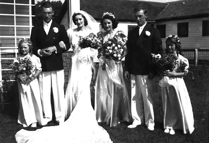 Six people in wedding attire pose outdoors with bouquets in front of a wooden house.