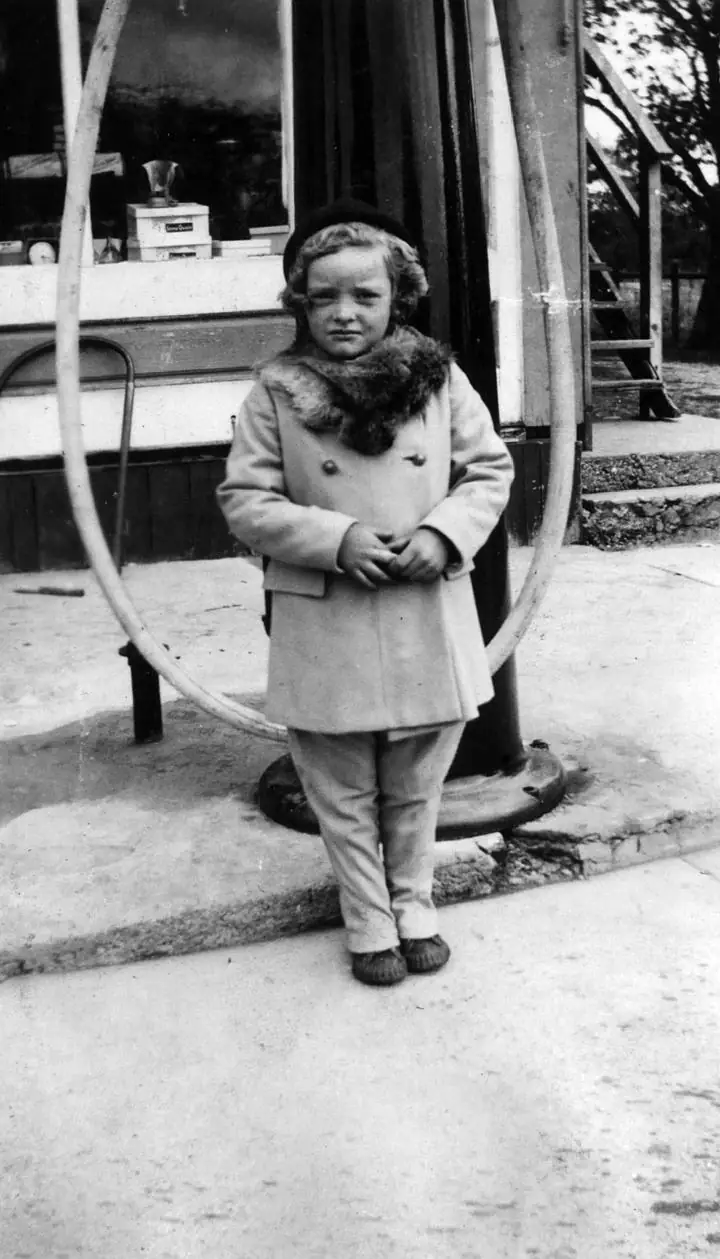 Child in a fur-collared coat standing beneath a circular frame outside a building.