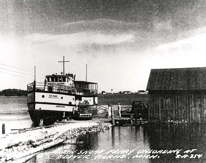 A ferry marked “NORTH SHORE” is moored beside a rocky pier, with vehicles and people near a large wooden shed.
