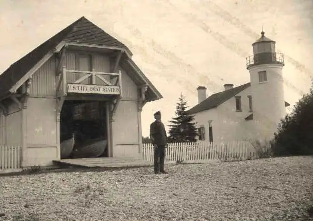 Sepia photograph of a lifeboat station, lighthouse, open boat bay, and a standing person.