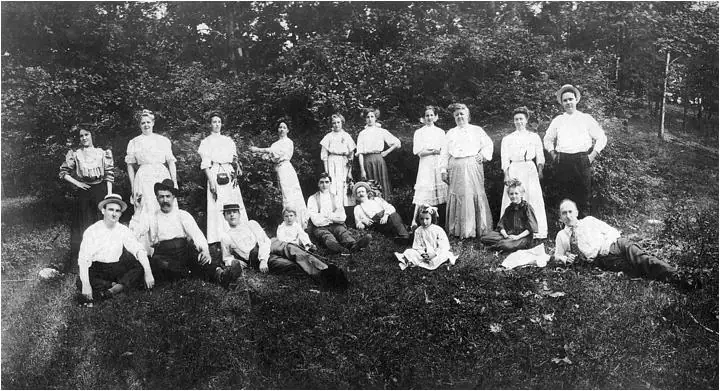 A large group of adults and children poses in a grassy clearing before a wooded backdrop.