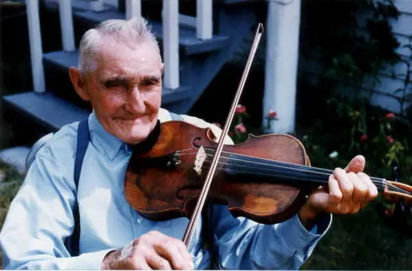An older man plays a violin in front of a porch and garden.