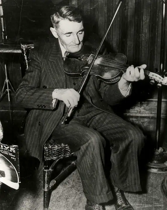 A seated man in a pinstriped suit plays a violin in a wood-paneled room.