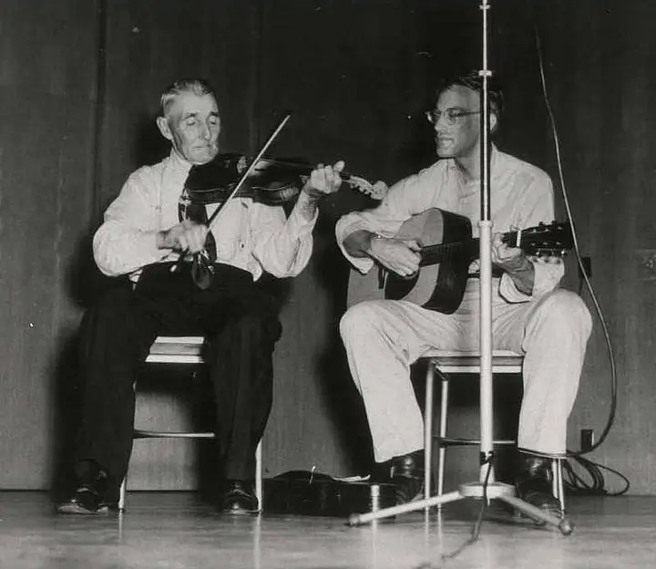 Two seated musicians perform with a violin and acoustic guitar beside a microphone stand.