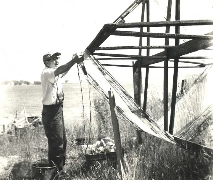 A man works with a large fishing net stretched across a wooden outdoor frame.